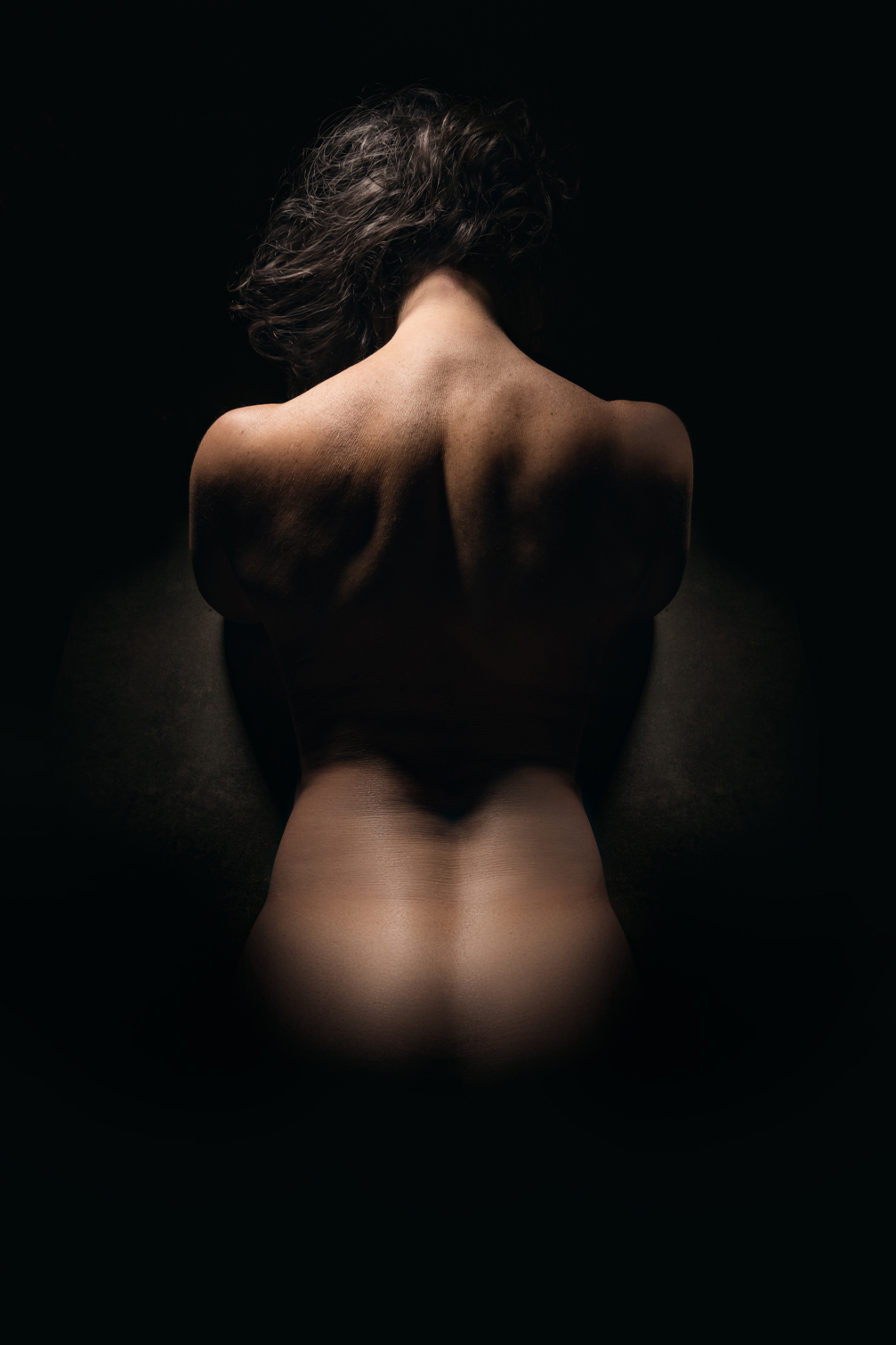 Fotografía de espaldas de mujer desnuda, entre las sombras, iluminada tenuemente resaltando sus curvas.